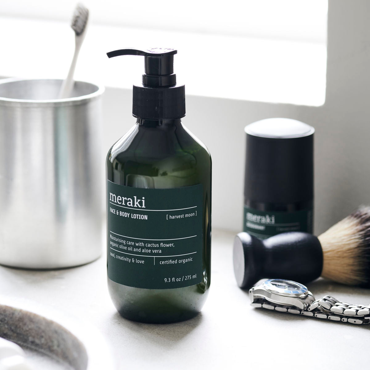 A bottle of Meraki Face & Body Lotion Organic, Harvest Moon with a pump sits on a bathroom counter near a toothbrush, shaving brush, deodorant, and wristwatch. This lotion is ECOCERT Cosmos certified for gentle daily care.