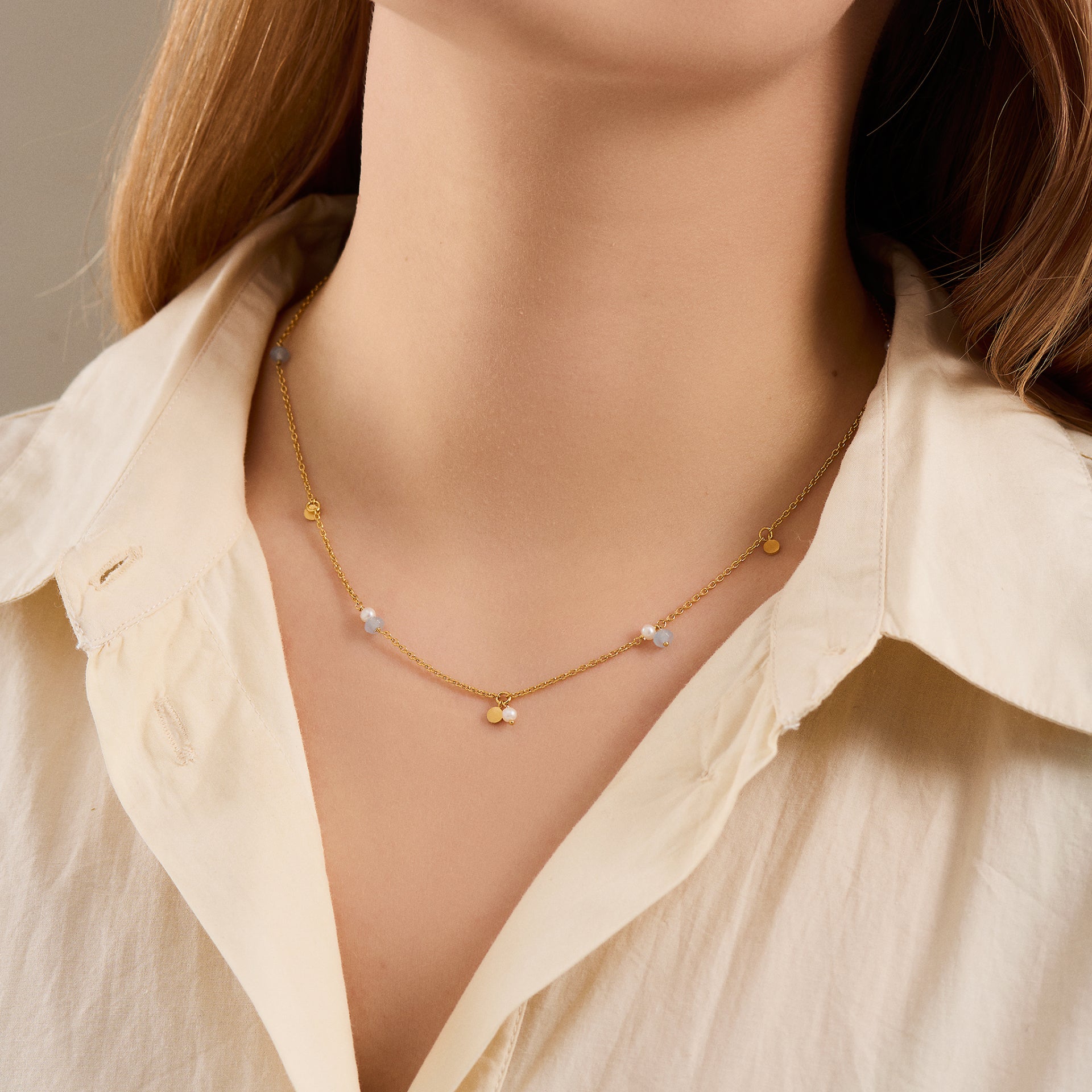 A person wearing a cream button-up shirt and the Pernille Corydon Afterglow Sea Necklace in silver with freshwater pearls and blue agate.