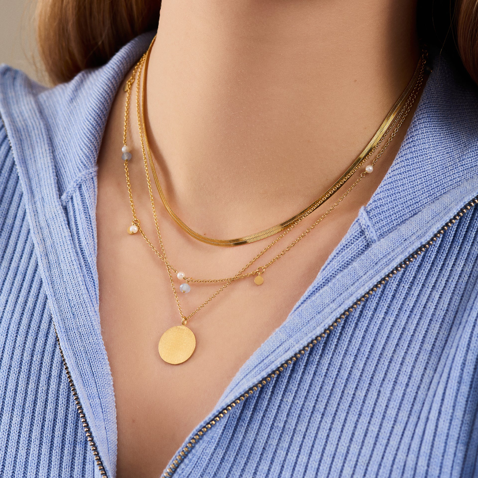 Close-up of a person wearing a light blue ribbed zip-up top and the Pernille Corydon Afterglow Sea Necklace in Silver with Freshwater Pearls and Blue Agate, echoing natural treasures.
