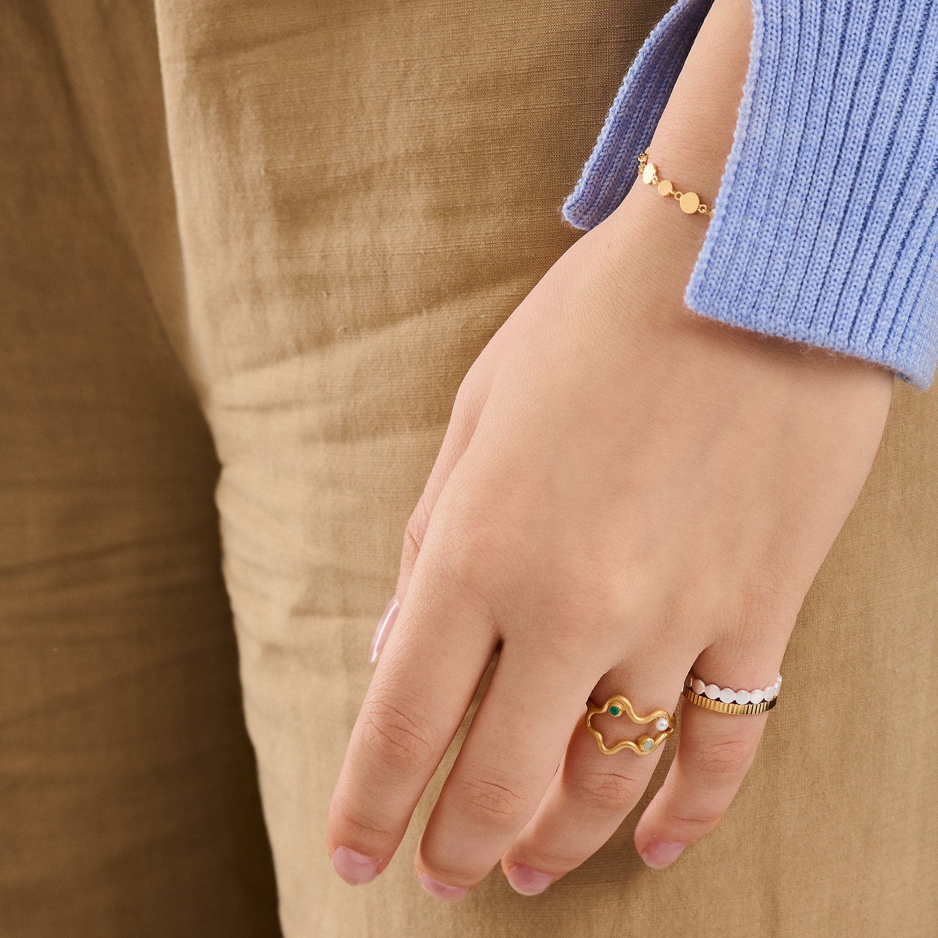 A person in a light blue long-sleeve shirt and beige pants showcases the matte Catena Ring in Gold by Pernille Corydon and a delicate gold bracelet on their left hand.