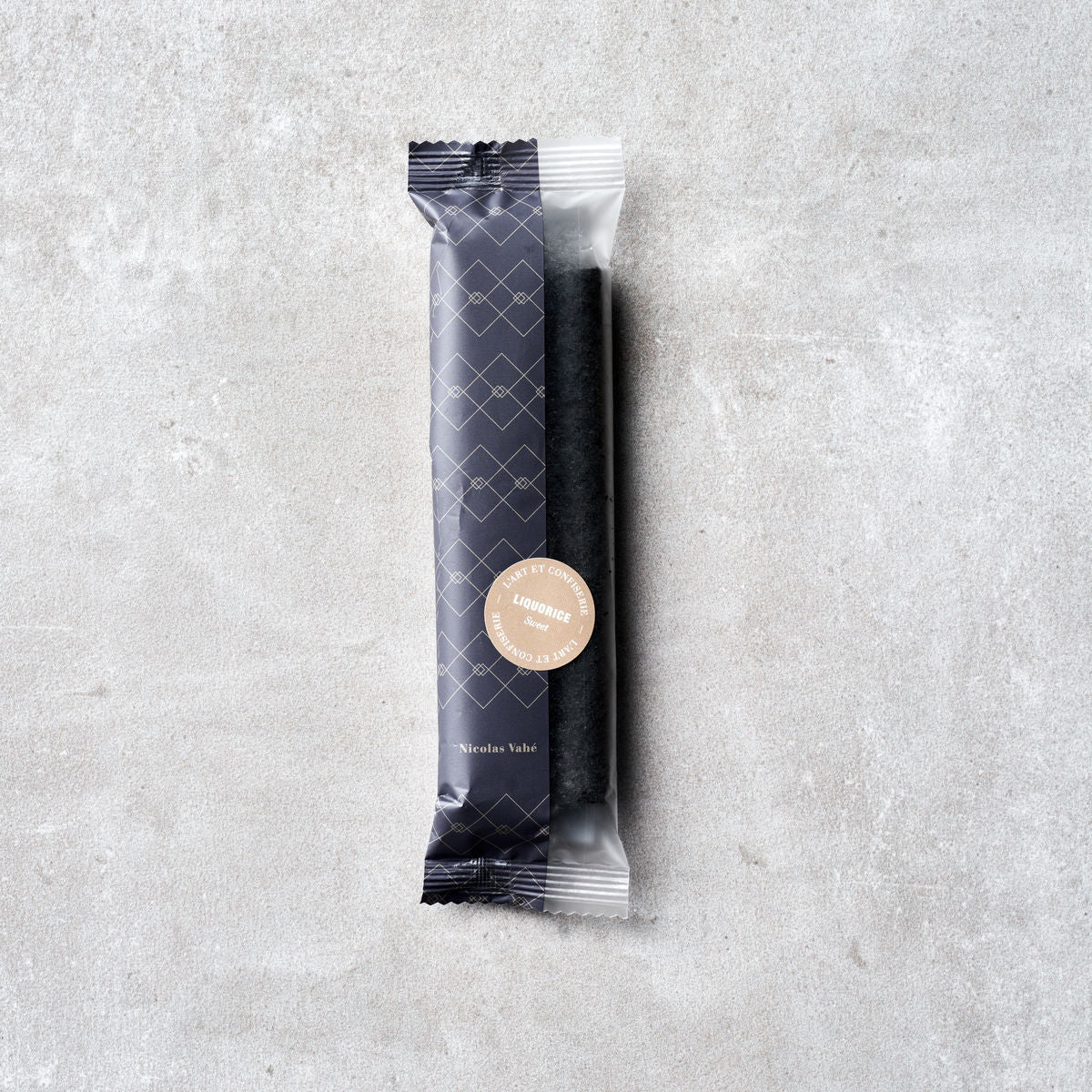 A single Sweet Soft Liquorice stick by Nicolas Vahé, in a black patterned wrapper with a beige label, is placed on a light gray textured surface—perfect as a stylish gift.