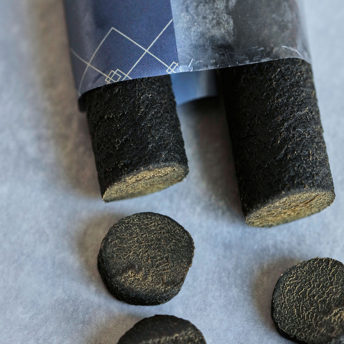 Two cylindrical Sweet Soft Liquorice sticks by Nicolas Vahé, partly wrapped in blue paper, with two round licorice slices on a gray surface—an ideal gift for any sweets lover.