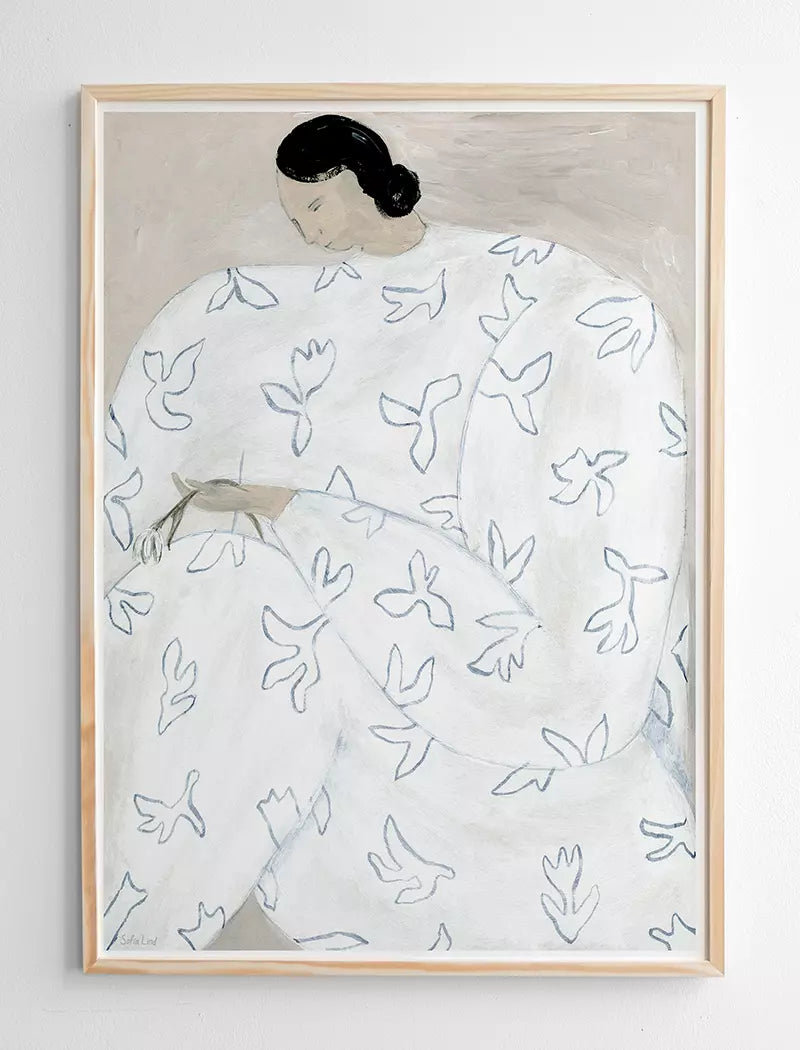 White Flower Print 50x70 cm by Fine Little Day features a seated figure in a white garment with blue doves, head bowed and arms on lap, framed and displayed against a white wall.