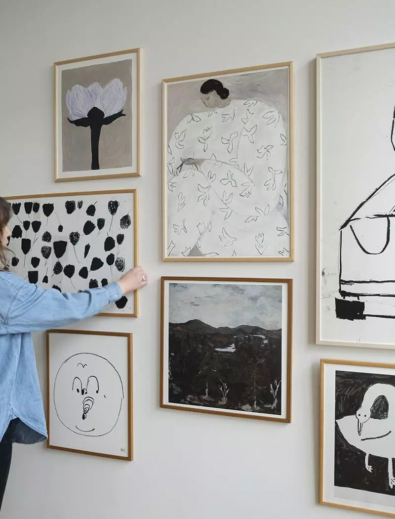 A person arranges a gallery wall featuring the Fine Little Day White Flower Print (50x70 cm) among seven framed abstract and minimalist pieces, inspired by Swedish artist Sofia Lind, on a light-colored wall.