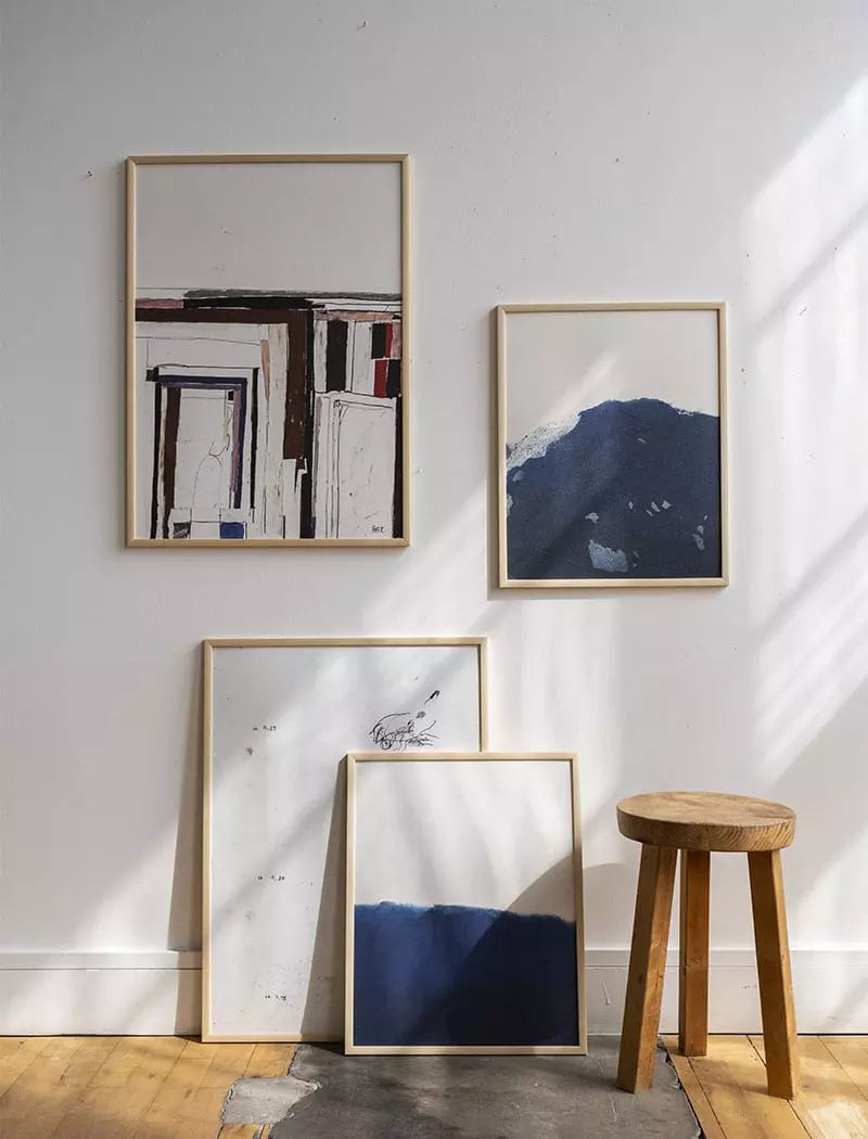 Two Ocean 1 Prints (40x50 cm) by Fine Little Day hang on a white wall, while two more lean on the floor beside a wooden stool. Sunlight casts shadows across this limited edition abstract display.