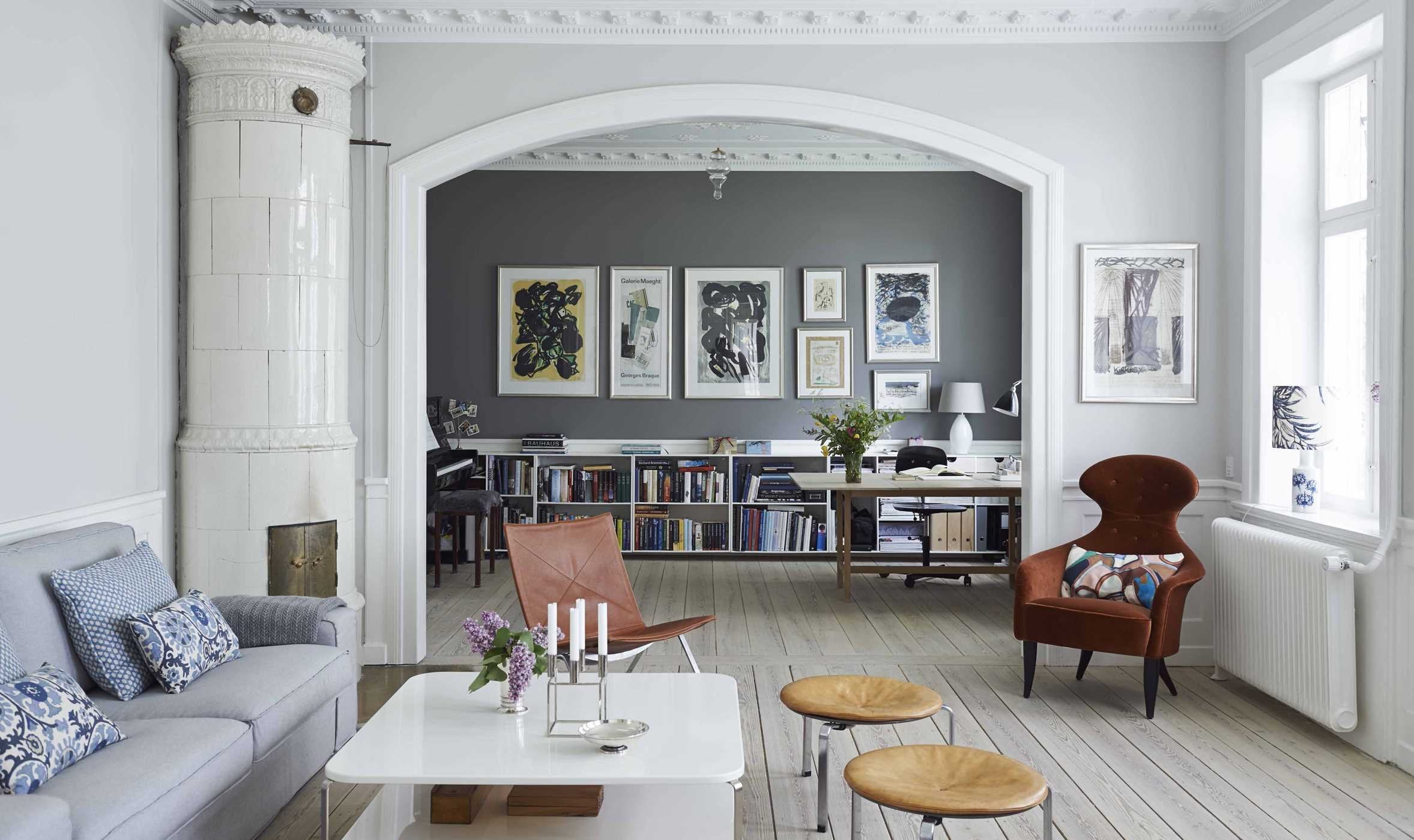 A modern living room with light gray walls and a white stove, connecting to a home office with bookshelves, captures the elegant style found in The Scandinavian Home - Interiors Inspired by Light by Niki Brantmark.