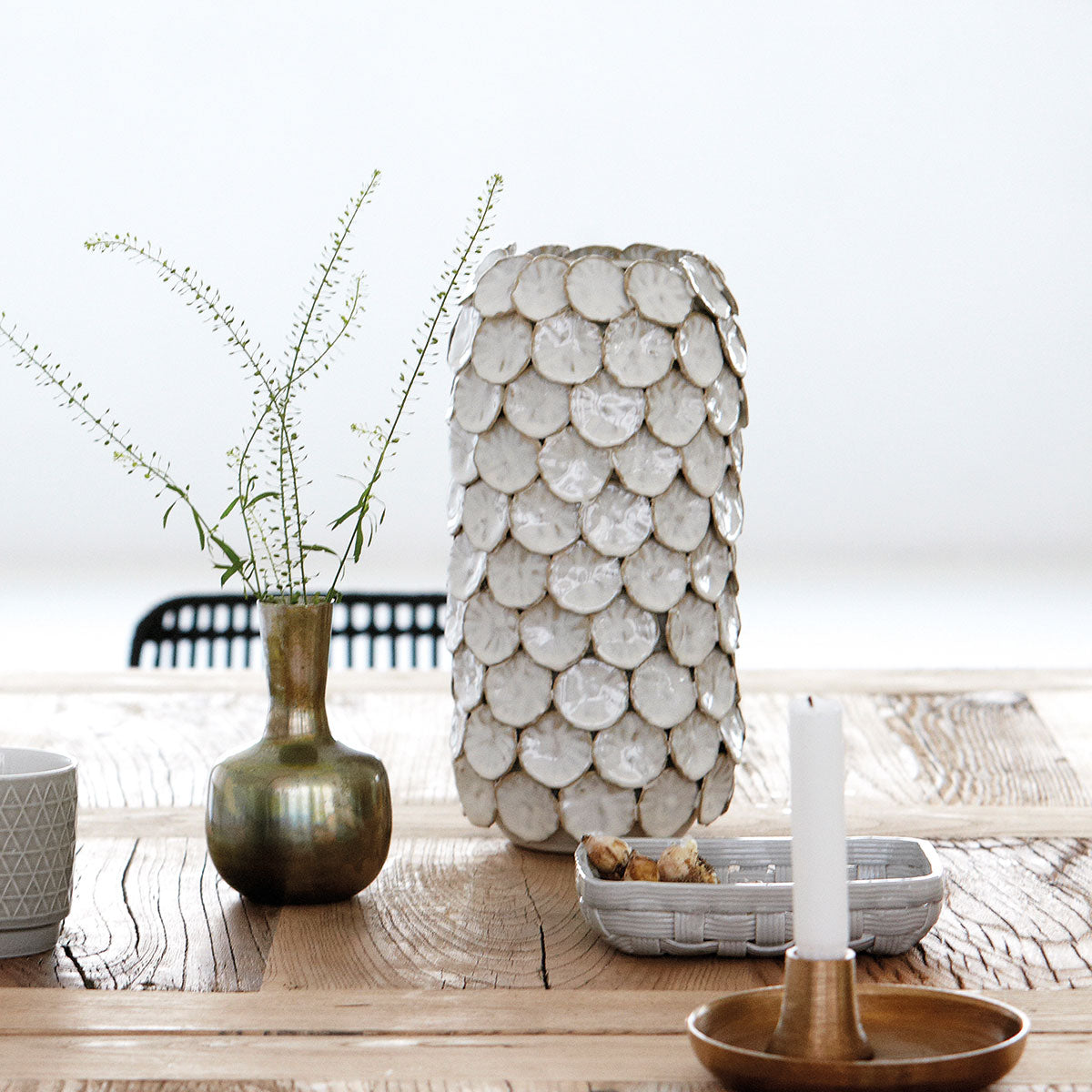 A wooden table with a House Doctor Vase Dot Ceramic 30cm, a small green vase with sprigs, a white candle in a wooden holder, and a bowl of cookies.