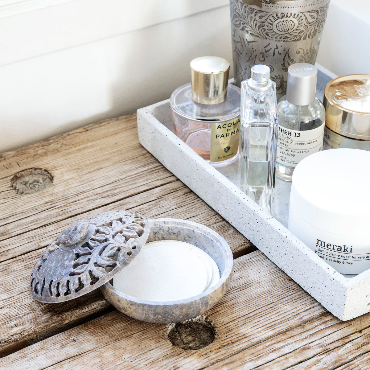 A reusable Meraki Facial Cleansing Sponge rests on a rustic wooden surface next to a tray of Meraki skincare and perfume bottles, adding style and convenience to your facecare routine.