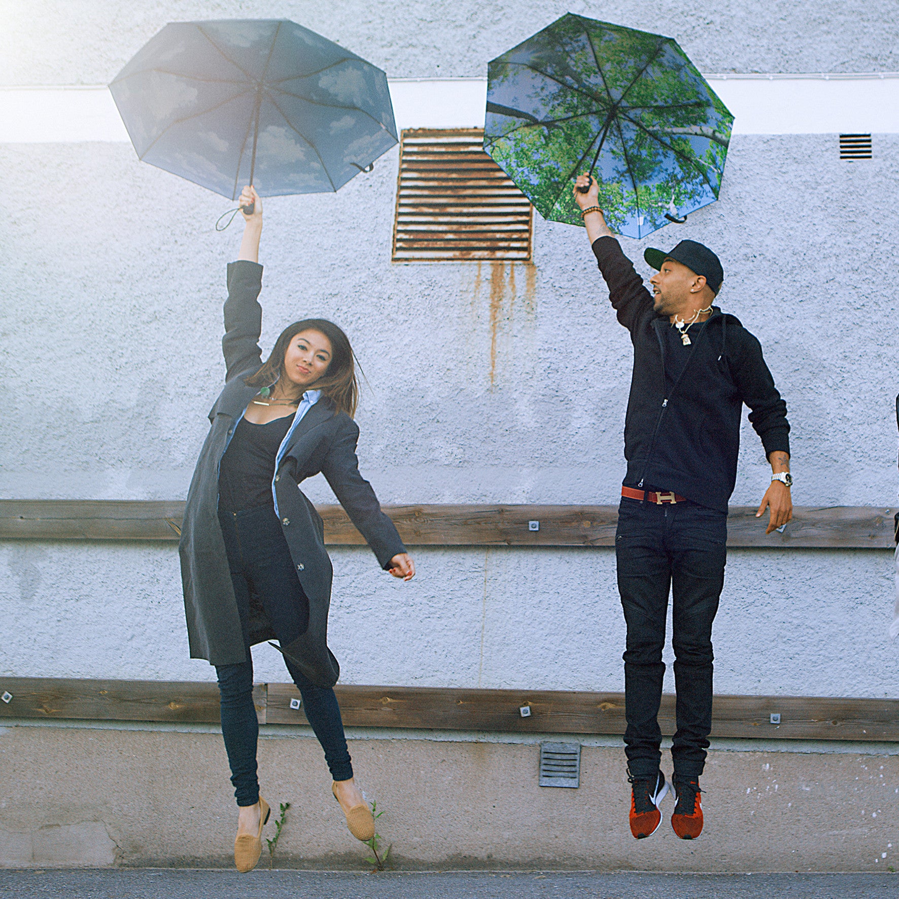 Two people with Happysweeds Umbrella Forest jump against a grey wall, looking as though the umbrellas are lifting them into the air.