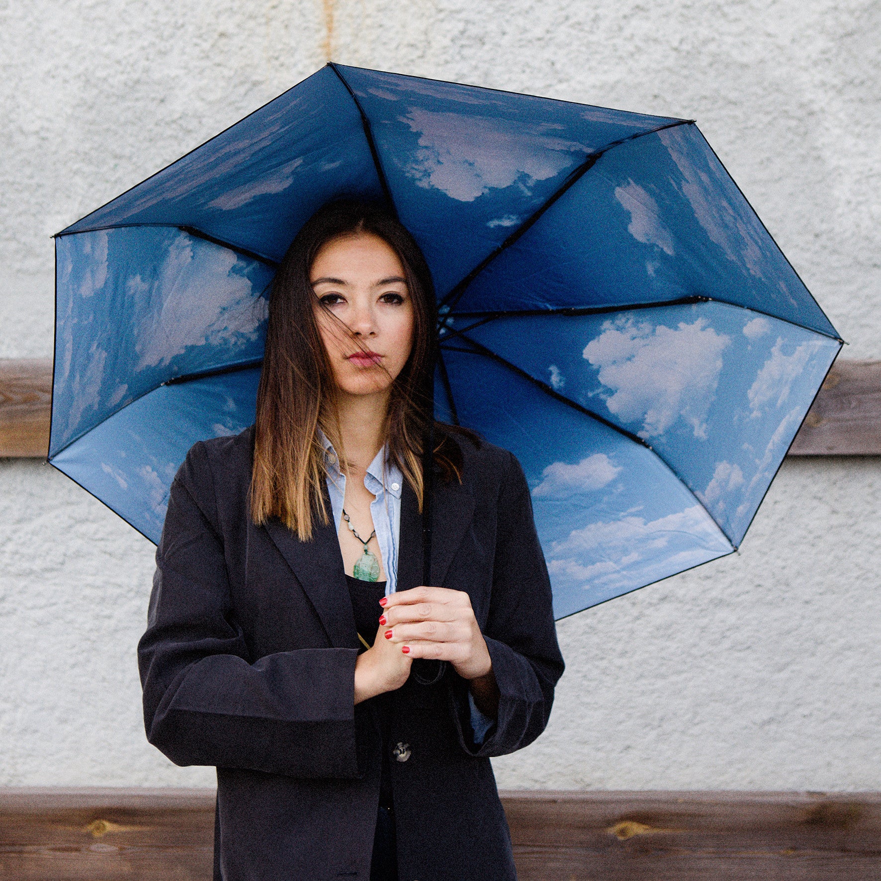 A woman in a dark blazer holds the Happysweeds Umbrella Sky Lake, featuring a sky and clouds-patterned interior, as she stands against a textured light wall.