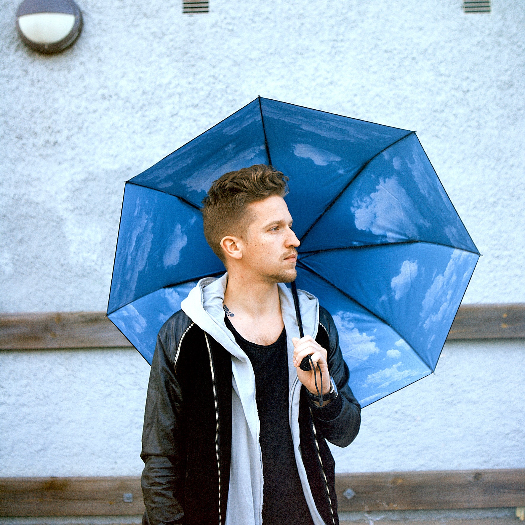 A man holds an open Happysweeds Umbrella Sky Lake, featuring a sky and clouds print, while standing against a light-colored wall.