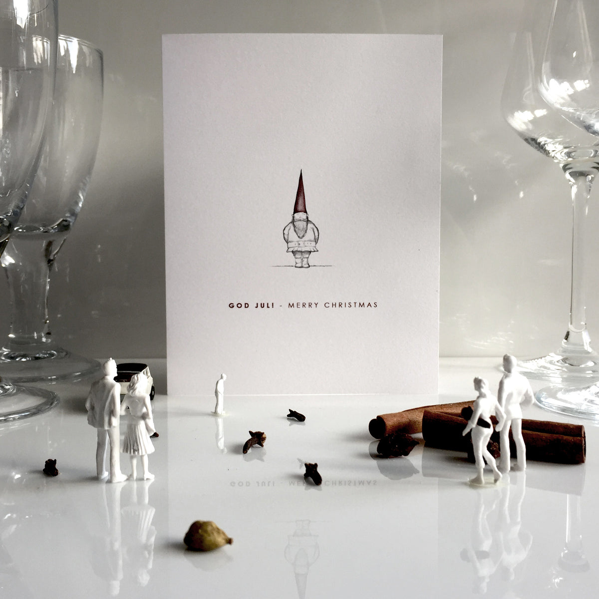 A holiday card with a drawing of a gnome and the words God Jul - Merry Christmas stands among miniature white figurines, glassware, spices, and cinnamon sticks on a white surface.