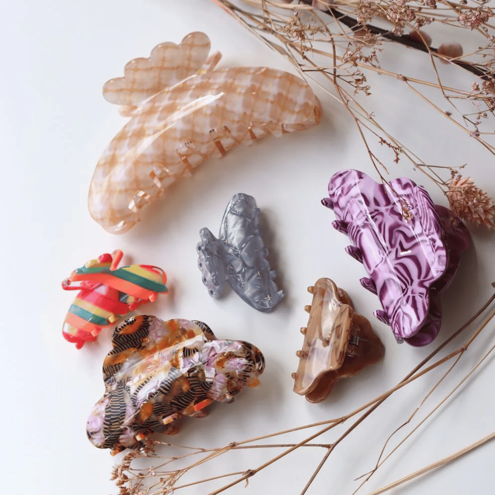 Six colorful, patterned hair claw clips of various shapes and sizes are arranged on a white surface, surrounded by delicate dried plants.