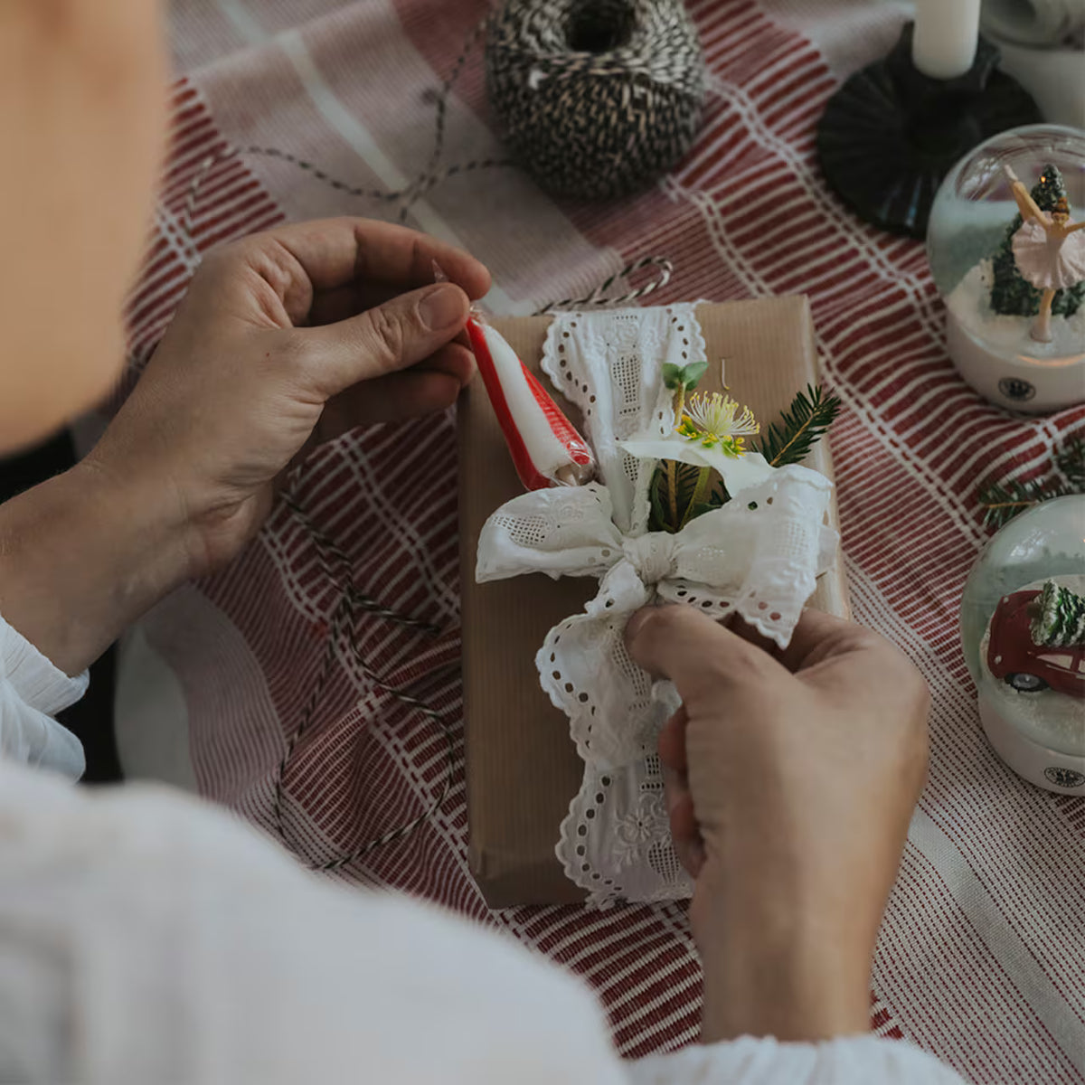 A person ties a white lace ribbon around a brown paper-wrapped gift, adorned with small flowers and greenery, on a red and white tablecloth with ornaments and a Strömshaga Umbrella Lollipop, Polka—made in Sweden—nearby.