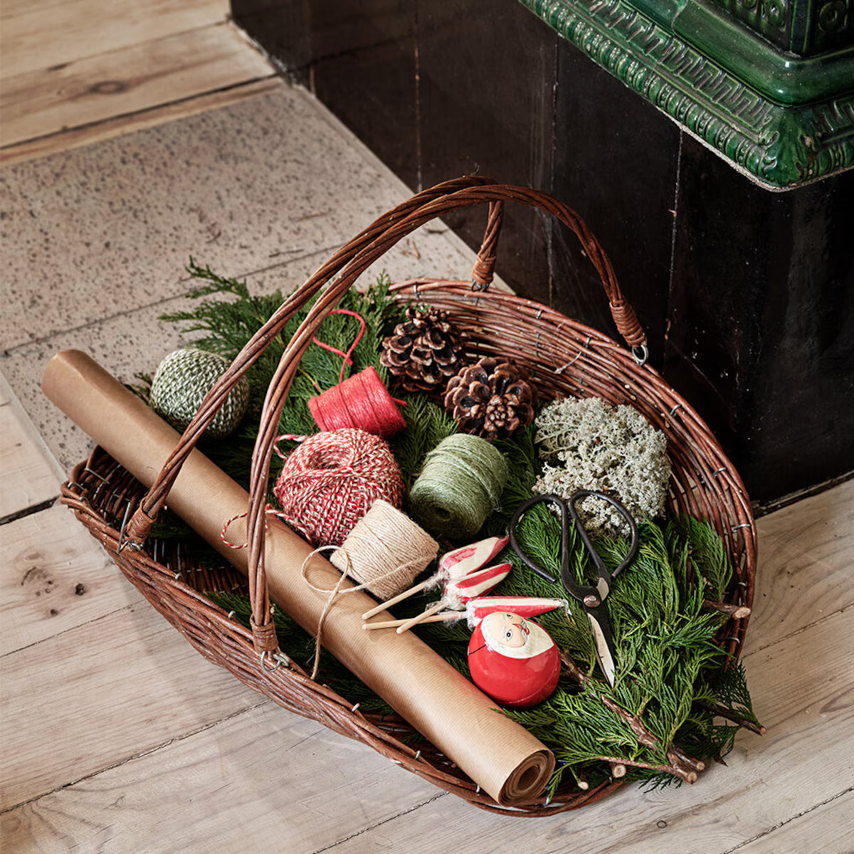 A wicker basket on a wooden floor holds pine branches, pinecones, twine, yarn, kraft paper, scissors, a spool of red ribbon, and decorative moss—featuring charming holiday or gift-wrapping supplies that evoke Scandinavian coziness. Nestled among the treasures is the Umbrella Lollipop by Strömshaga (Polka), made in Sweden, adding a playful and festive touch to the scene.