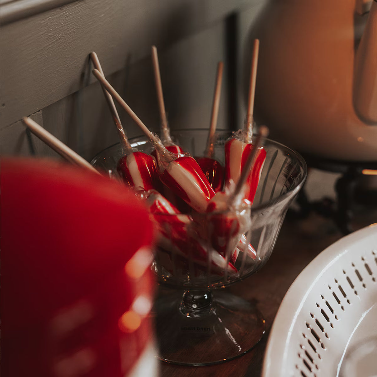 A glass bowl holds Strömshaga Umbrella Lollipop, Polka—red and white striped mint lollipops with sticks—resting on a wooden surface beside a white dish and a red object, all softly lit indoors; these classic Swedish treats are crafted with care.