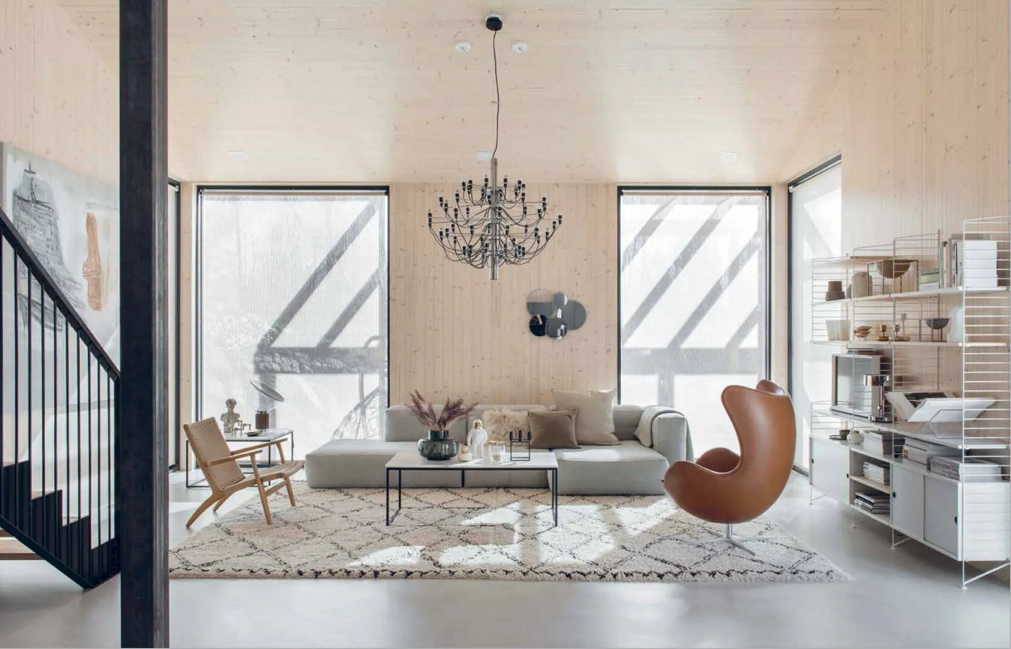 Inspired by Nordic Interior Book from Cozy Publishing, this modern living room features light wood walls, large windows, a gray sofa, brown egg chair, wooden armchair, patterned rug, and minimalist décor filled with natural light.