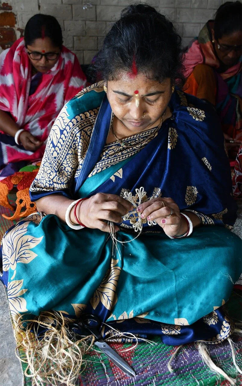 A woman in a blue sari sits on the floor meticulously weaving fibers to craft a delicate Nisse Santa Ornament Handmade in Jute, Red and Off-white by Strömshaga, while other women work alongside her in the background. They are focused on creating these charming rustic Christmas decorations together in their workshop or community space.