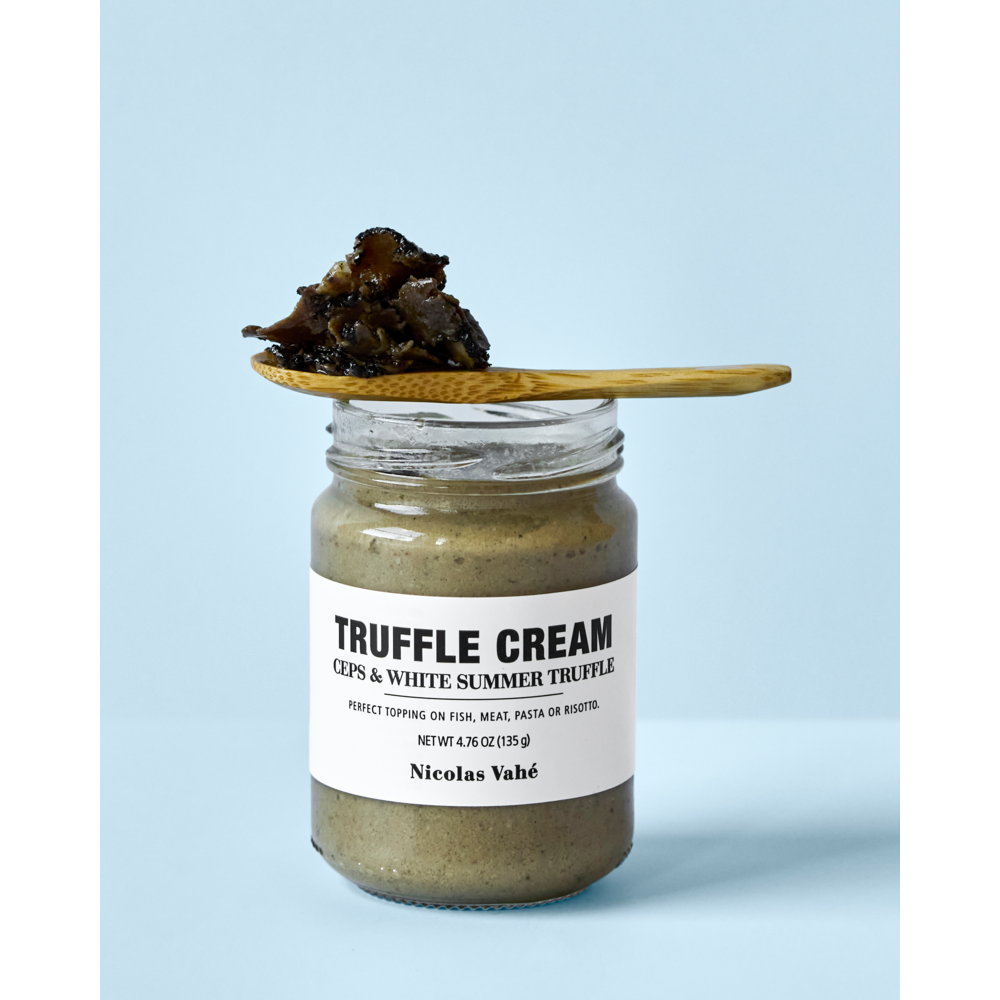 A jar of Nicolas Vahé Truffle Cream, Ceps & White Summer Truffle rests on a light blue surface, with a wooden spoon holding a rich mixture of white summer truffle and porcini mushrooms balanced across the jar’s open top.