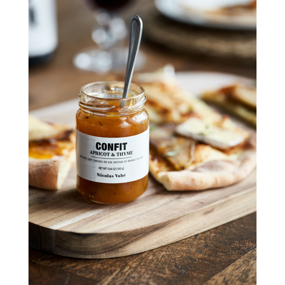 A jar of Nicolas Vahé’s Confit, Apricot & Thyme with a spoon rests on a wooden board beside sliced flatbread—an elegant addition to any cheese platter or perfect cheese pairing for rustic entertaining.