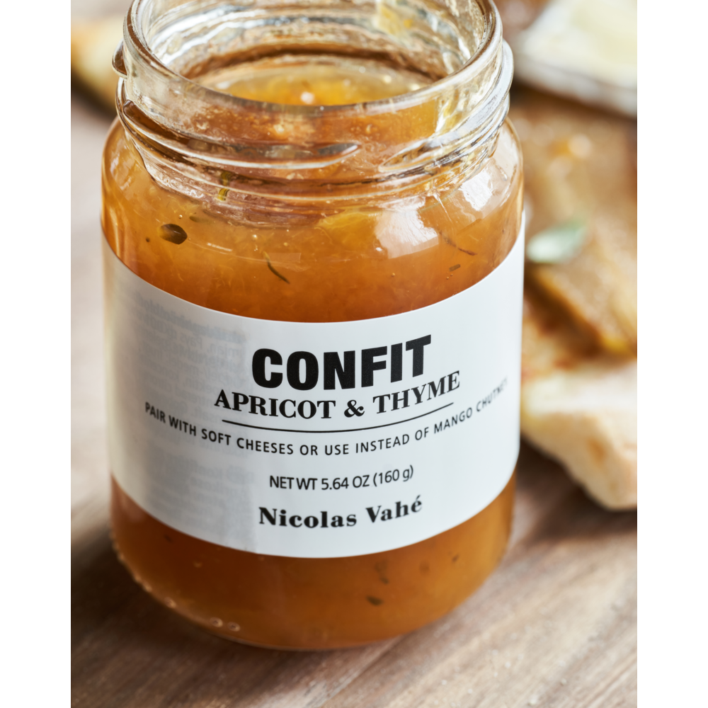 A jar of Nicolas Vahé Confit, Apricot & Thyme rests on a wooden surface, its label recommending it as an ideal cheese pairing. Blurred bread slices in the background hint at its place on your next cheese platter.