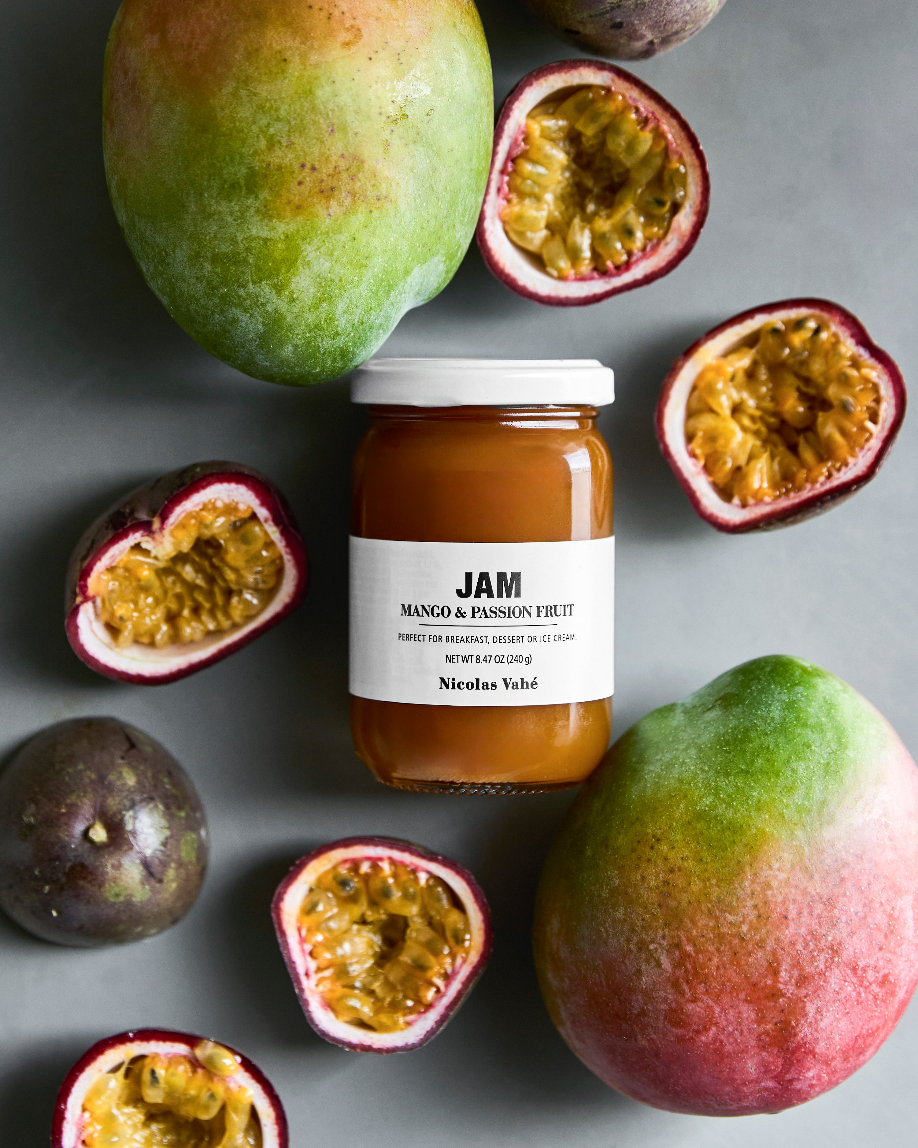 A jar of Jam, Mango & Passion Fruit from Nicolas Vahé is surrounded by whole and halved passion fruits and two whole mangoes on a gray surface. This delicious preserve from Nicolas Vahé beautifully captures the vibrant colors and flavors of tropical fruit.