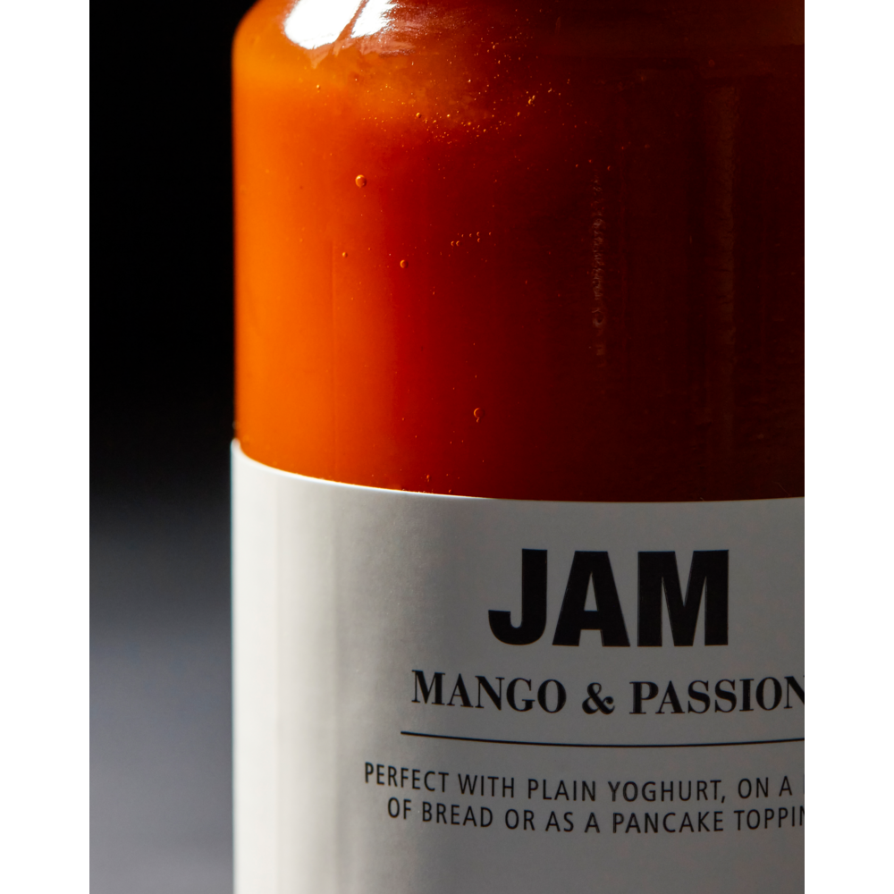 A close-up of a glass jar filled with vibrant orange fruit jam labeled Jam, Mango & Passion Fruit. The label from Nicolas Vahé suggests enjoying this delicious mango and passion fruit jam with plain yogurt, on bread, or as a topping for pancakes. The dark, blurred background highlights the inviting appeal of this Nicolas Vahé creation.