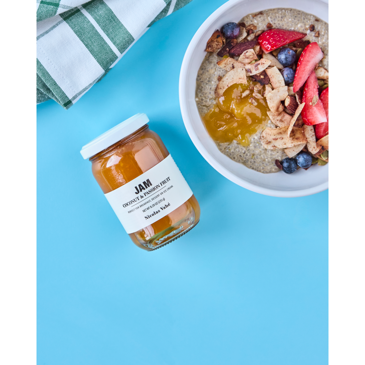 A jar of Nicolas Vahé Jam, Coconut & Passion Fruit sits beside a bowl of oatmeal topped with strawberries, blueberries, granola, and the same jam on a blue surface. A green and white towel is nearby.