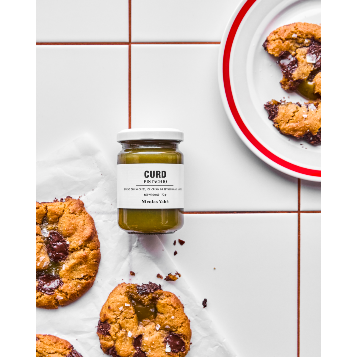 A jar of Nicolas Vahé Curd, Pistachio rests on white tile beside chocolate chip cookies, some filled with curd. In the upper right corner, a red-rimmed plate holds pieces of cookie.