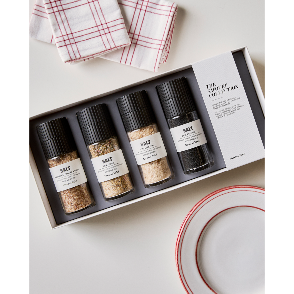 A boxed set of four Nicolas Vahé salt grinders, labeled "Gift Box Savoury Salts" and featuring Parmesan, Tomato & Basil Salt, Black Salt, Wild Garlic Salt, and Chili Salt in a savory collection gift box with a ceramic grinder, sits next to a red and white checkered towel and a white plate with a red rim on a white surface.