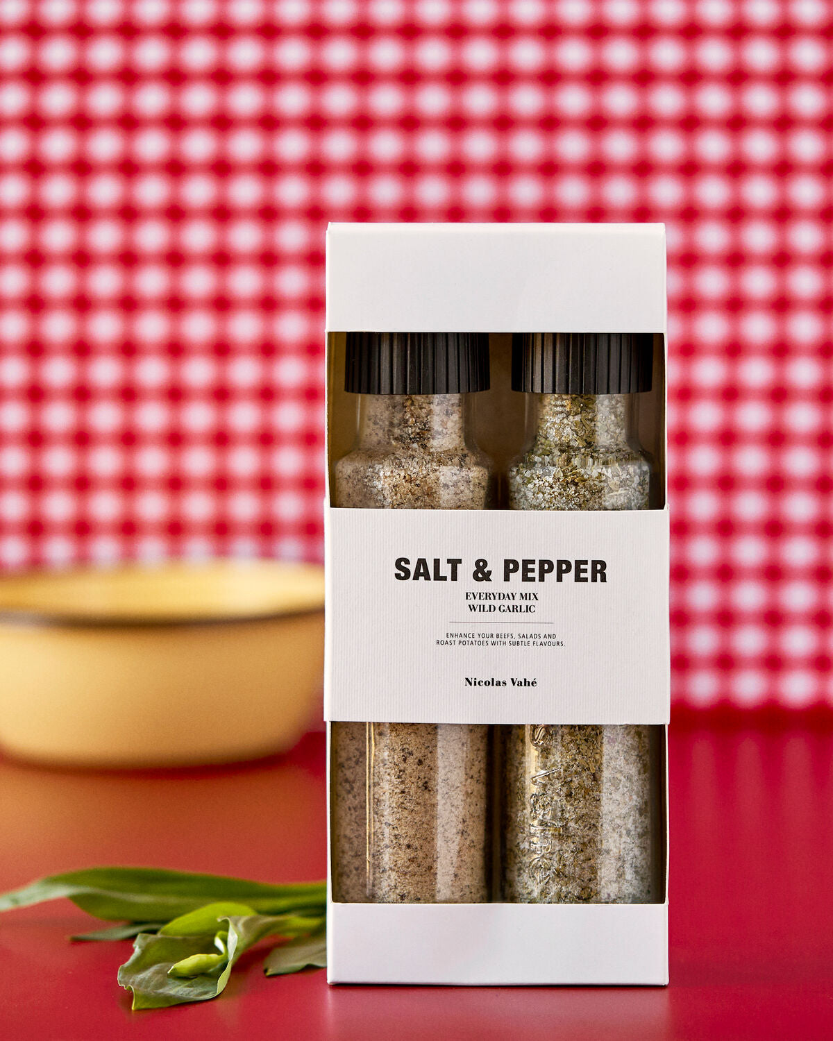 Two glass grinders, one filled with Everyday Mix Salt & Pepper and the other with Wild Garlic Salt by Nicolas Vahé, are packaged together in a sleek white gift box labeled “Gift Box, Everyday Mix Salt & Pepper and Wild Garlic Salt.” A yellow bowl and fresh green leaves are arranged nearby against a red and white checkered background. This Nicolas Vahé set is ideal for enhancing your favorite dishes with the flavors of wild garlic salt or an everyday salt and pepper blend.