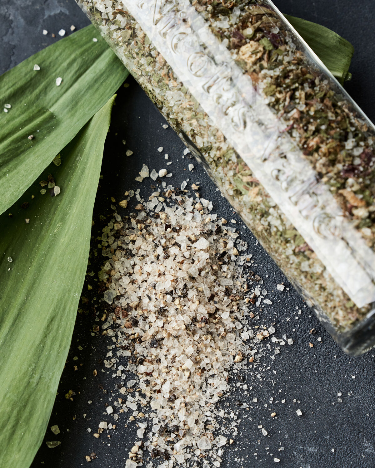 A glass jar from the Nicolas Vahé Gift Box—Everyday Mix Salt & Pepper and Wild Garlic Salt—spills onto a dark surface, with fresh green leaves nearby. The coarse salt blend combines aromatic dried herbs and spices, creating a flavorful mix ideal for enhancing any dish.