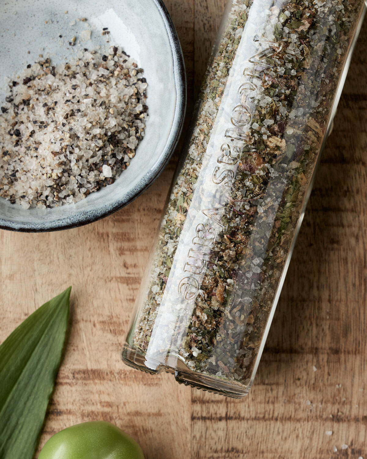 A close-up of Nicolas Vahé’s Wild Garlic Salt from the Gift Box, Everyday Mix Salt & Pepper and Wild Garlic Salt, displayed in a bowl and a glass container on a wooden surface beside a green leaf and fruit.