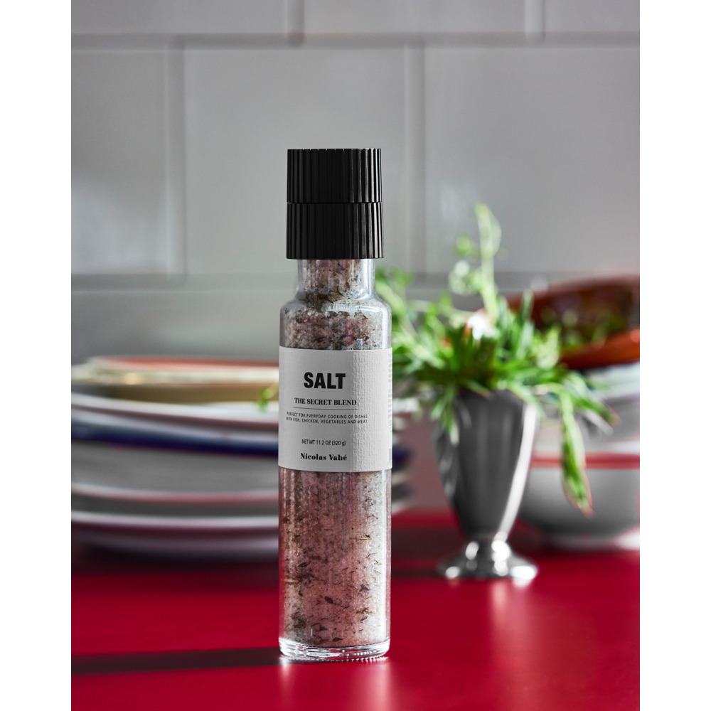 A glass salt grinder labeled "Salt, The Secret Blend" from Nicolas Vahé sits on a red countertop, filled with premium sea salt, with stacked plates and a small vase of aromatic herbs in the background, against a white tiled wall.