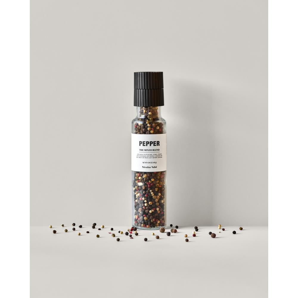 A clear pepper grinder by Nicolas Vahé, filled with white, red, green, pink, and black peppercorns (Pepper, The Mixed Blend), stands upright on a light surface with scattered peppercorns.