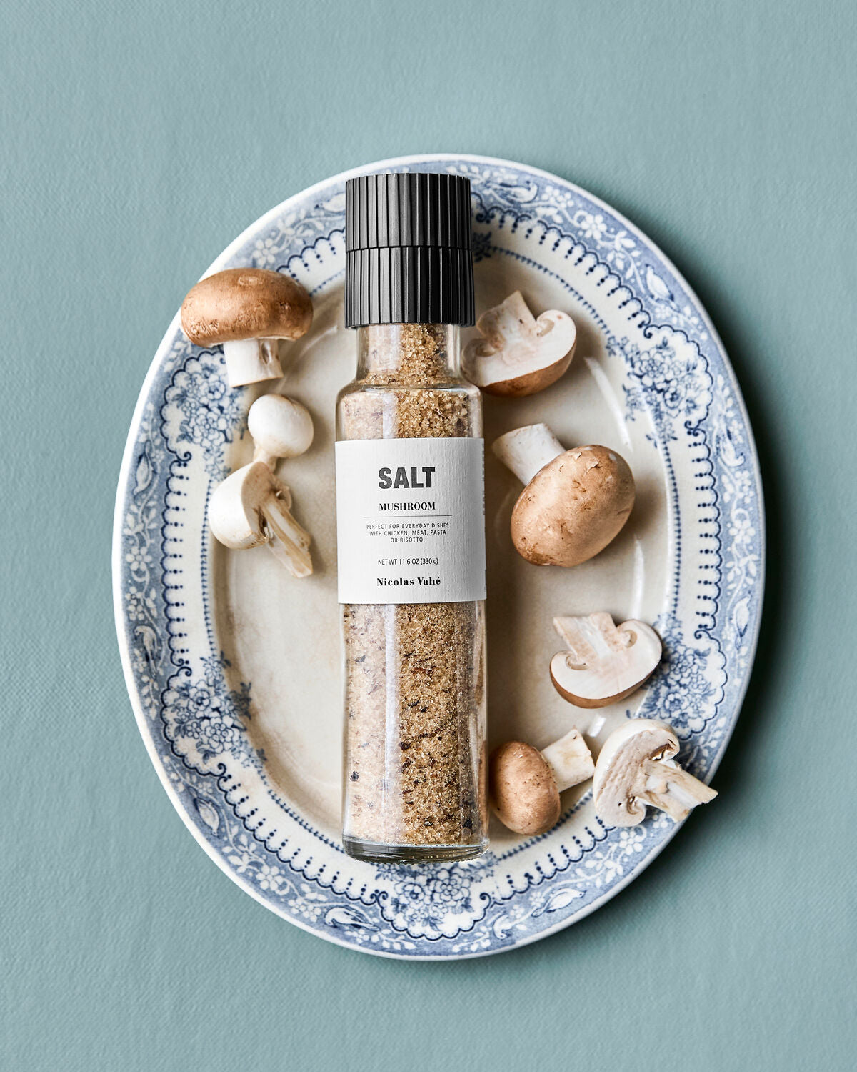 A bottle of Nicolas Vahé Salt, Mushroom with a ceramic grinder is displayed on a blue and white decorative plate, surrounded by whole and sliced brown and white mushrooms against a light blue background.