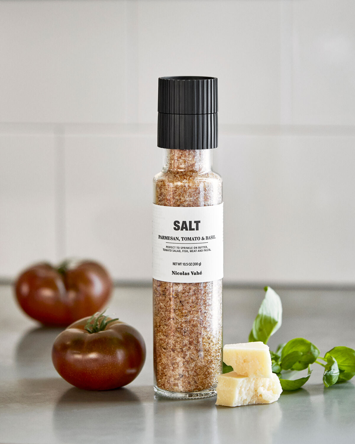 A bottle of Nicolas Vahé Salt, Parmesan, Tomato & Basil stands on a countertop, surrounded by two tomatoes, fresh basil leaves, and cubes of parmesan cheese; the backdrop is a tiled kitchen wall.