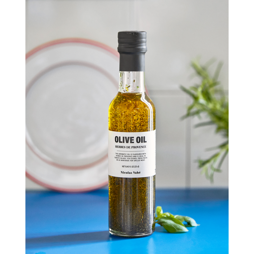 A clear glass bottle of Nicolas Vahé Olive Oil w. Herbes de Provence, 25 cl, sits on a blue countertop. The label reads “OLIVE OIL HERBES DE PROVENCE.” In the background, a white plate and fresh green herbs are softly blurred.