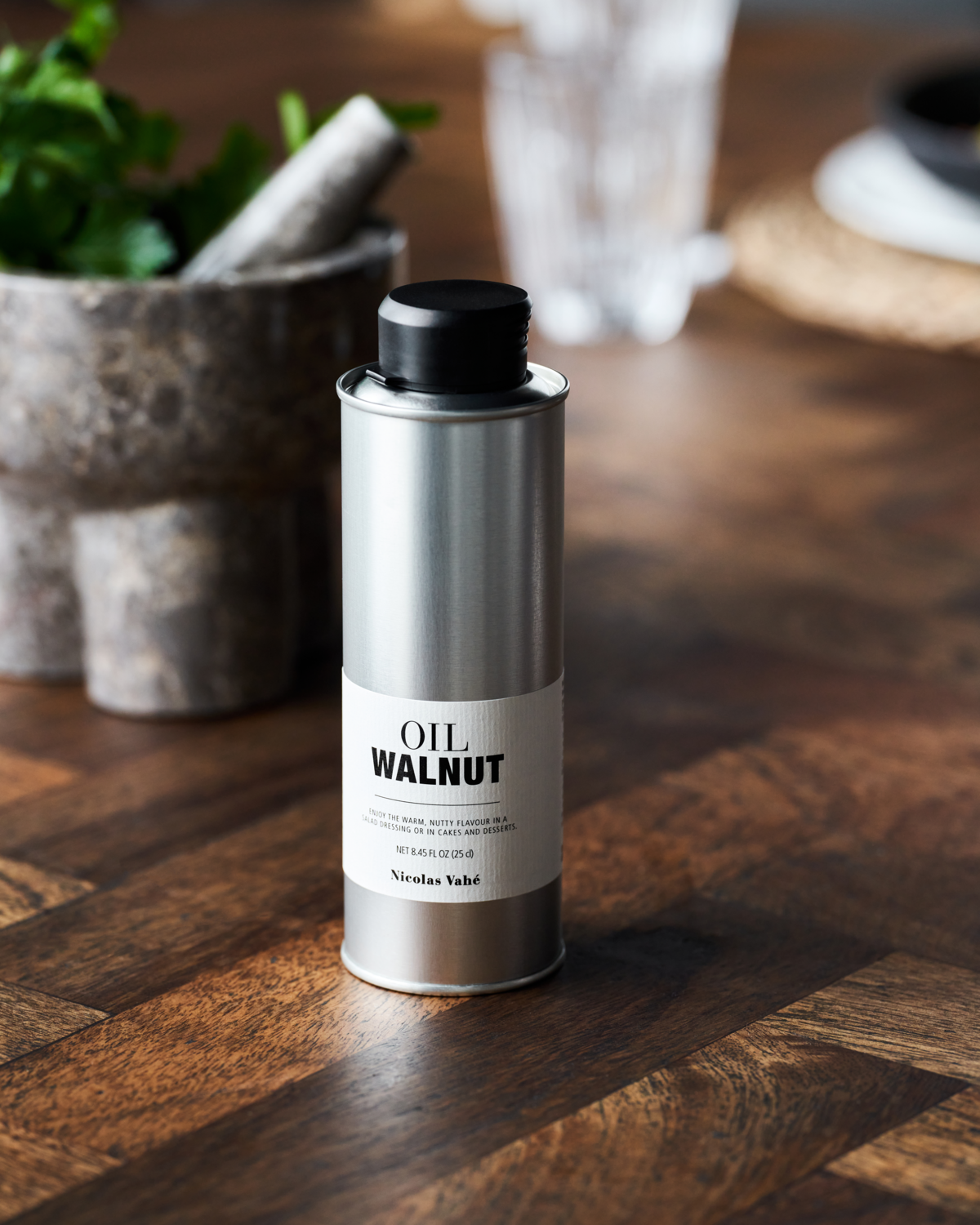 A metal canister labeled "Walnut Oil" by Nicolas Vahé, topped with a black cap, rests on a wooden surface, highlighting its premium quality French walnut oil with kitchen items softly blurred in the background.