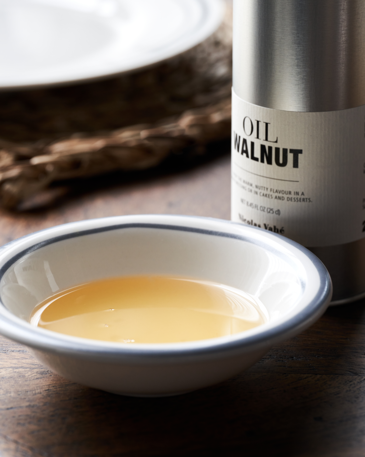 A small white bowl filled with light golden Walnut Oil sits on a wooden table beside a canister labeled Nicolas Vahé Walnut Oil, showcasing this premium French finishing oil.