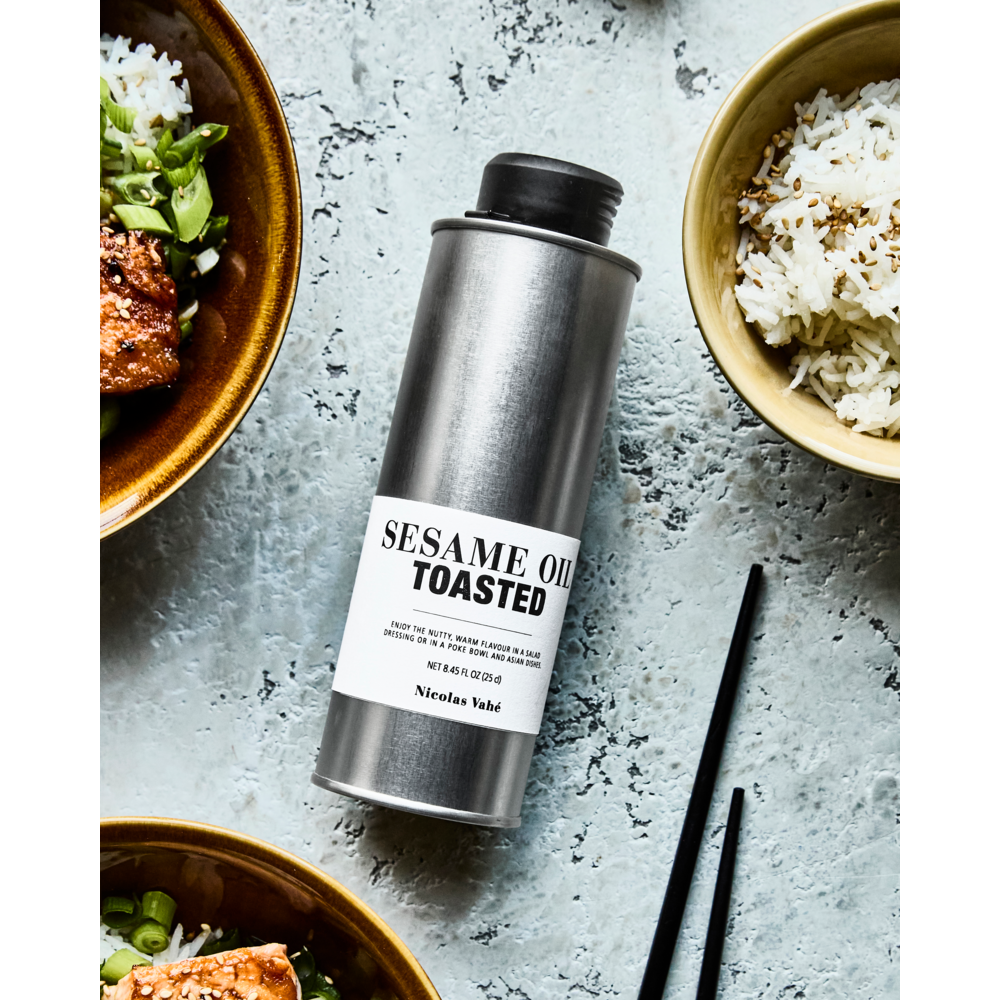 A metal can of Nicolas Vahé Toasted Sesame Oil is displayed on a white surface with bowls of rice, food topped with green onions, and black chopsticks—ideal for adding rich, nutty flavor to your favorite dishes.