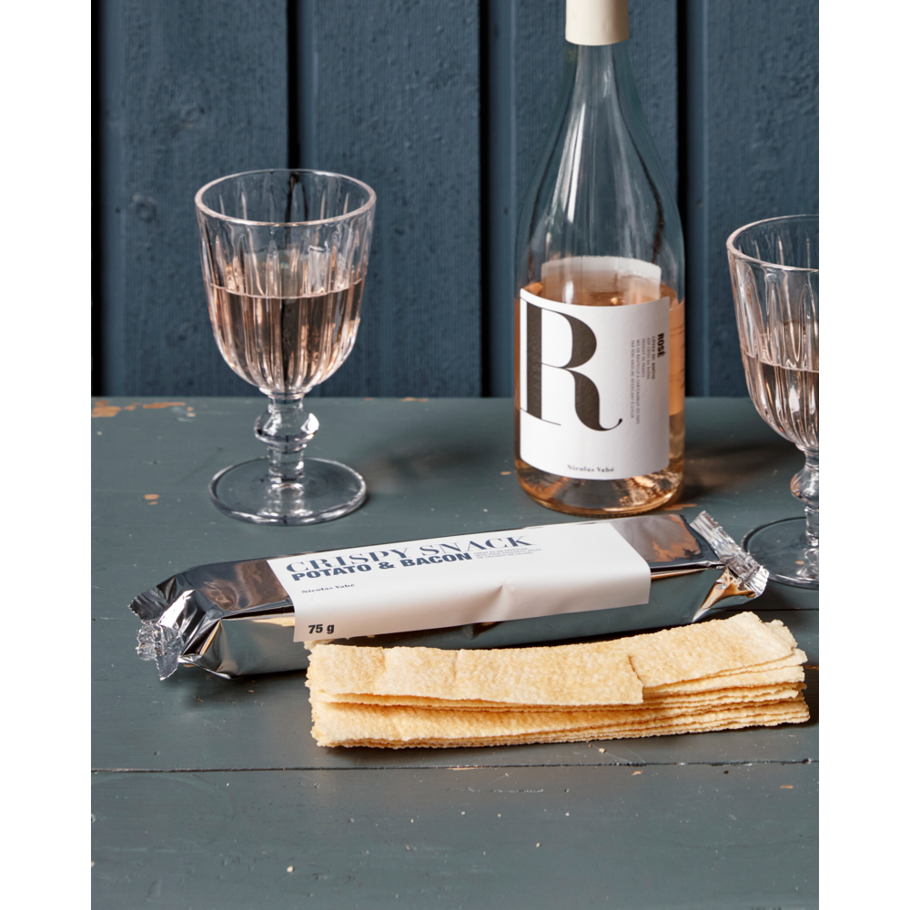 A bottle of rosé wine with a white label, two glasses of rosé, and a package of Nicolas Vahé Crispy Snack, Potato & Bacon, with stacked crisps, are arranged on a blue wooden table against a blue wooden wall.