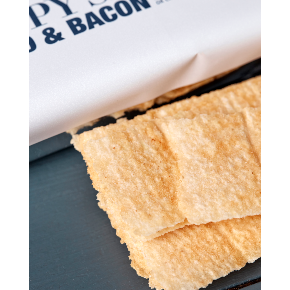 Close-up of rectangular, golden Crispy Snack, Potato & Bacon crackers by Nicolas Vahé stacked on a dark surface, with part of a white paper wrapper featuring blue text visible at the top.