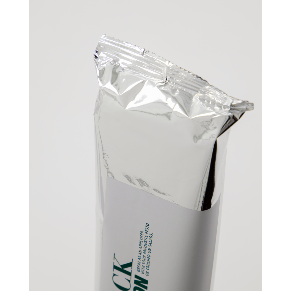 A partially opened silver foil wrapper on a white background reveals the top of a Nicolas Vahé Crispy Snack, Sour Cream & Onion, with part of its white label and dark green text visible—hinting at delicious long crisps inside.