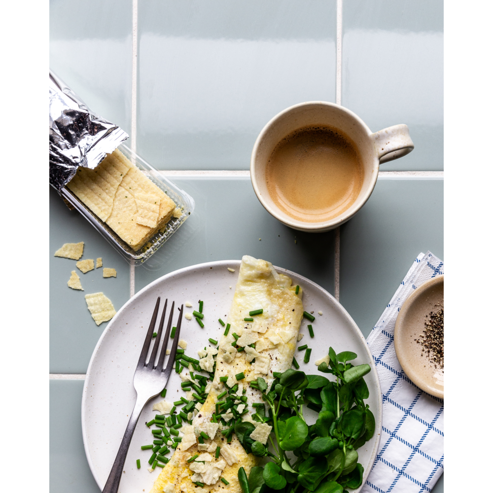 A plate with an omelette topped with chives and crackers, salad greens, a fork, coffee, a bowl of pepper, a blue-checked napkin, and Nicolas Vahé Crispy Snack, Sour Cream & Onion on a tiled surface.