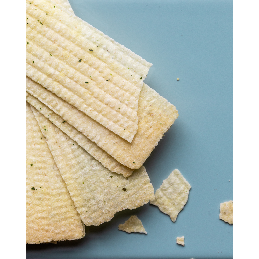 Rectangular, ridged Crispy Snack Sour Cream & Onion by Nicolas Vahé, fanned out on a light blue surface with a few broken pieces and crumbs nearby—a perfectly crispy potato treat.