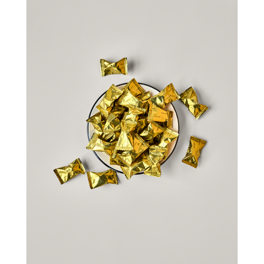 A bowl of Nicolas Vahé Cocoa Truffle, Caramel candies in shiny gold wrappers sits against a plain light background, with several caramel treats scattered around it.