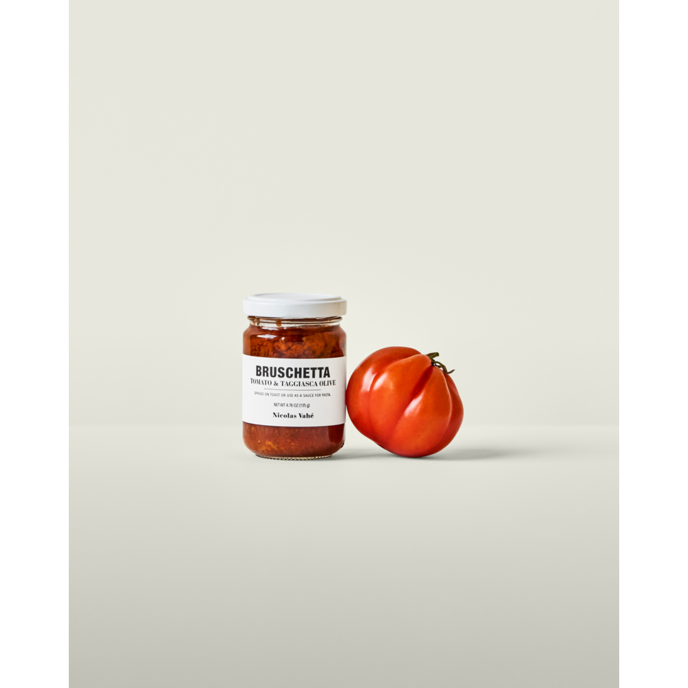 A jar of Nicolas Vahé Bruschetta, Tomato & Taggiasca Olives sits next to a ripe tomato on a light, plain surface with a neutral background.