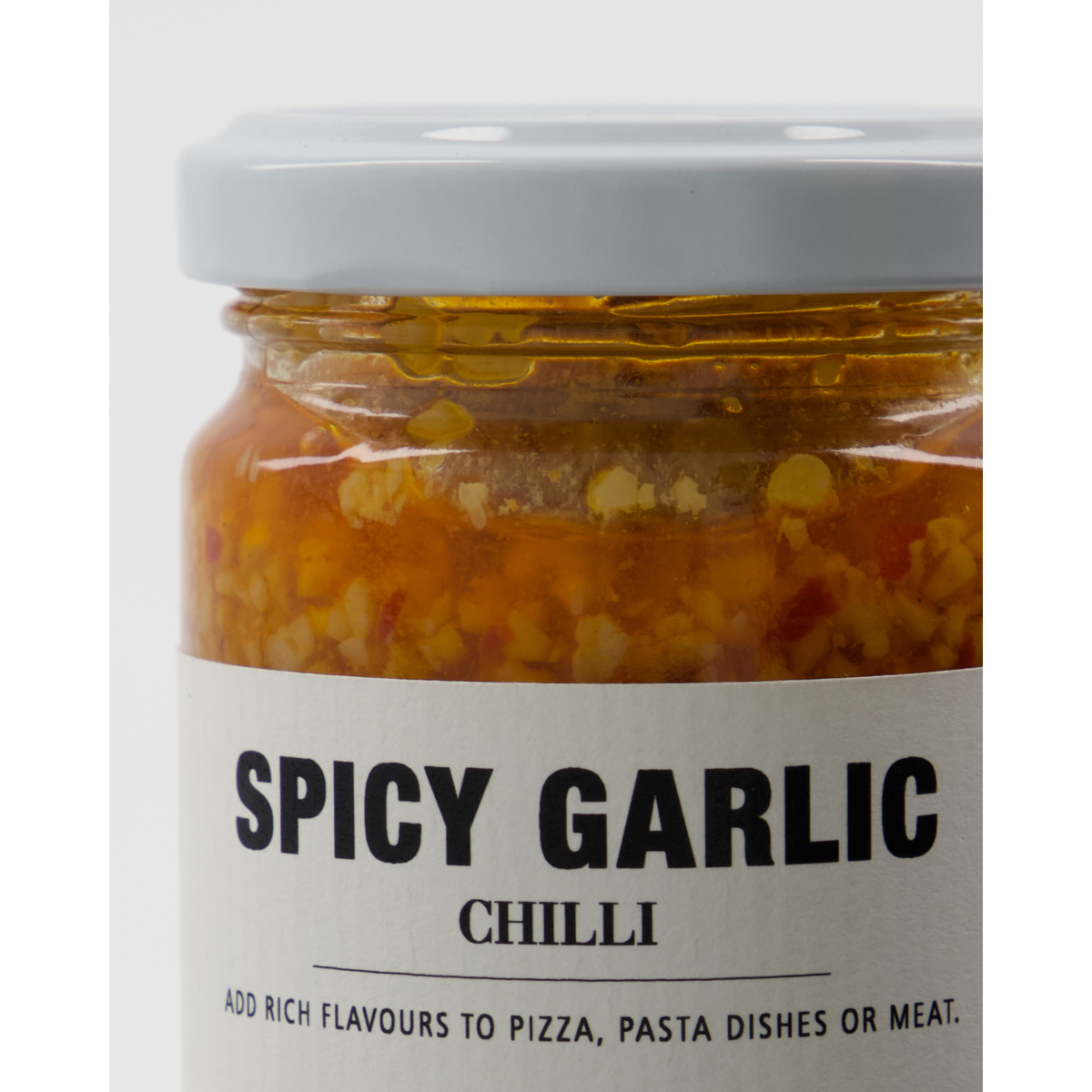 A close-up of a glass jar labeled Nicolas Vahé Spicy Garlic in Chilli Oil, showing chopped garlic and chili pieces. The label recommends adding this vibrant condiment to pizza, pasta, or meat for bold flavor.