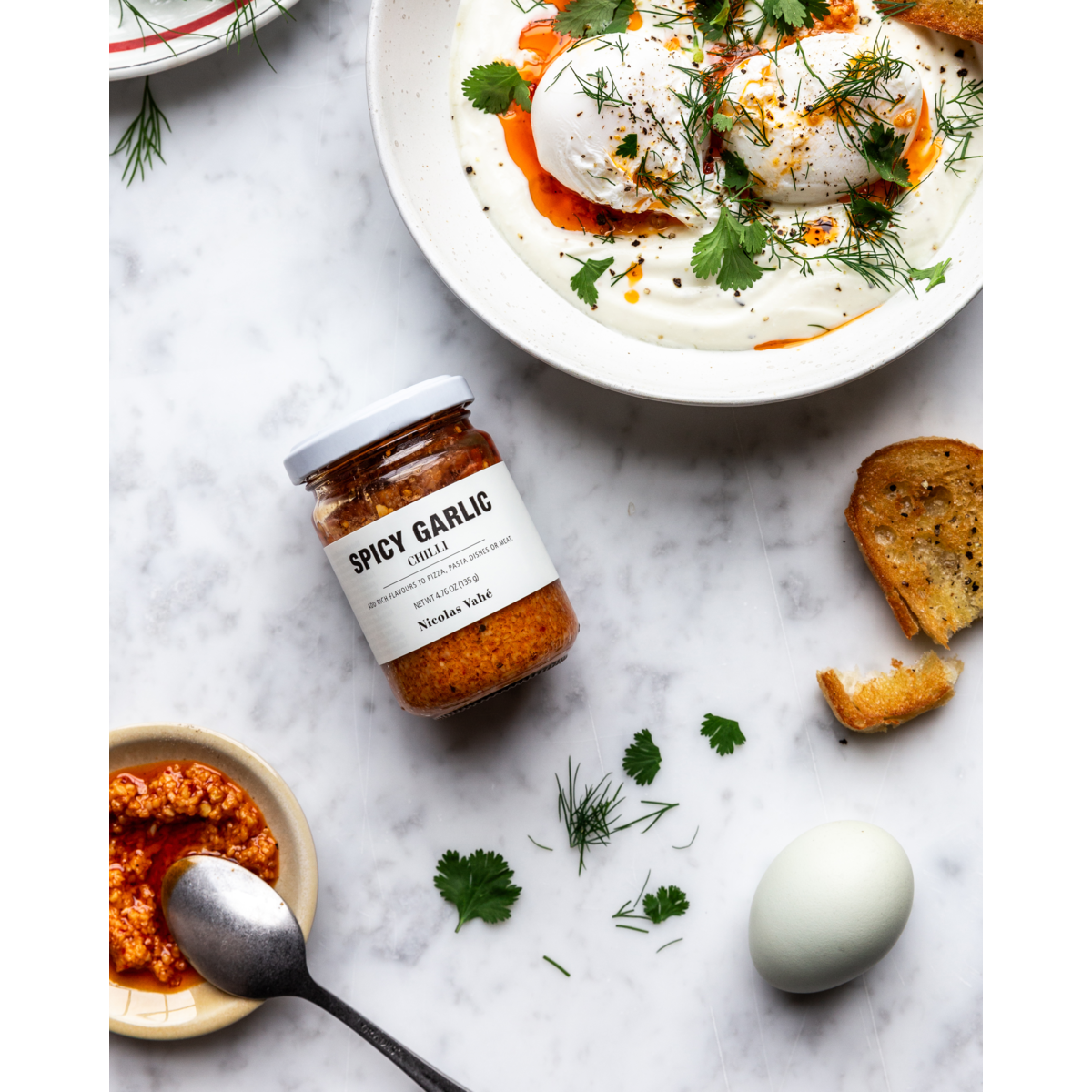 A jar of Nicolas Vahé Spicy Garlic in Chilli Oil sits on a white surface alongside a spoon, blue egg, toasted bread, scattered herbs, and a bowl of poached eggs on yogurt with herbs and aromatic chilli oil.